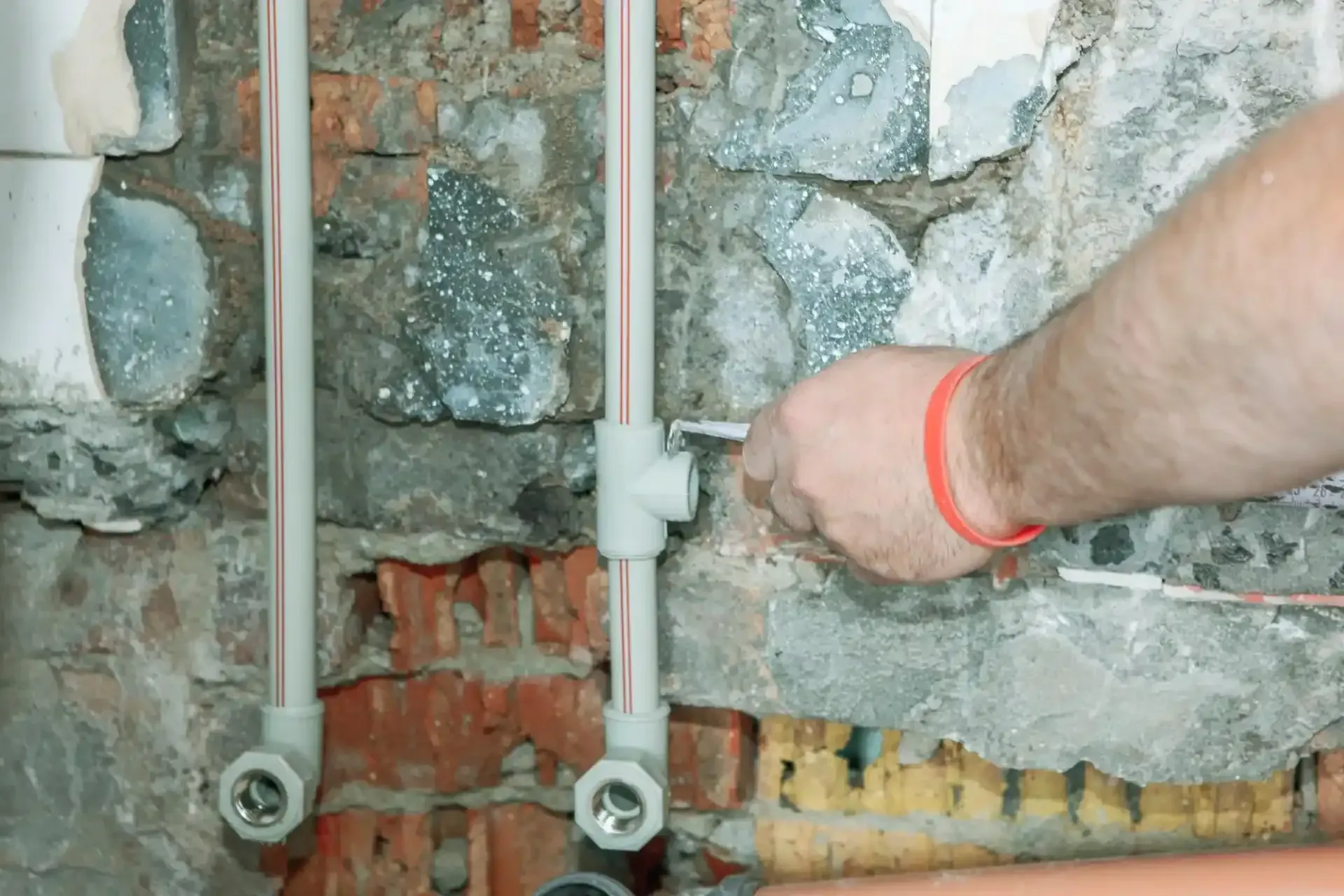 Idraulico che collega tubi di plastica bianca a un raccordo a T contro un muro di mattoni e pietra; è visibile un braccialetto rosso.