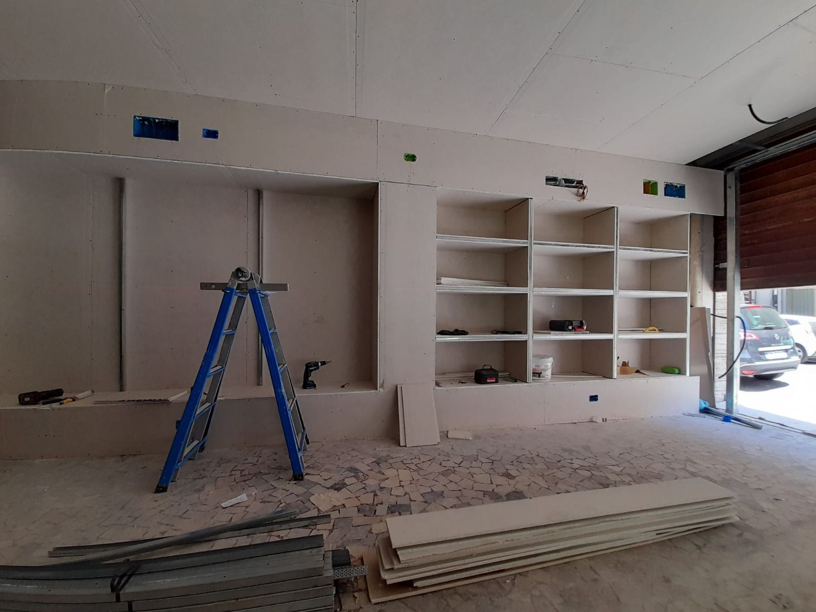 Interni in costruzione: scaffali in cartongesso, scala, attrezzi, muri parzialmente costruiti e porta del garage aperta.