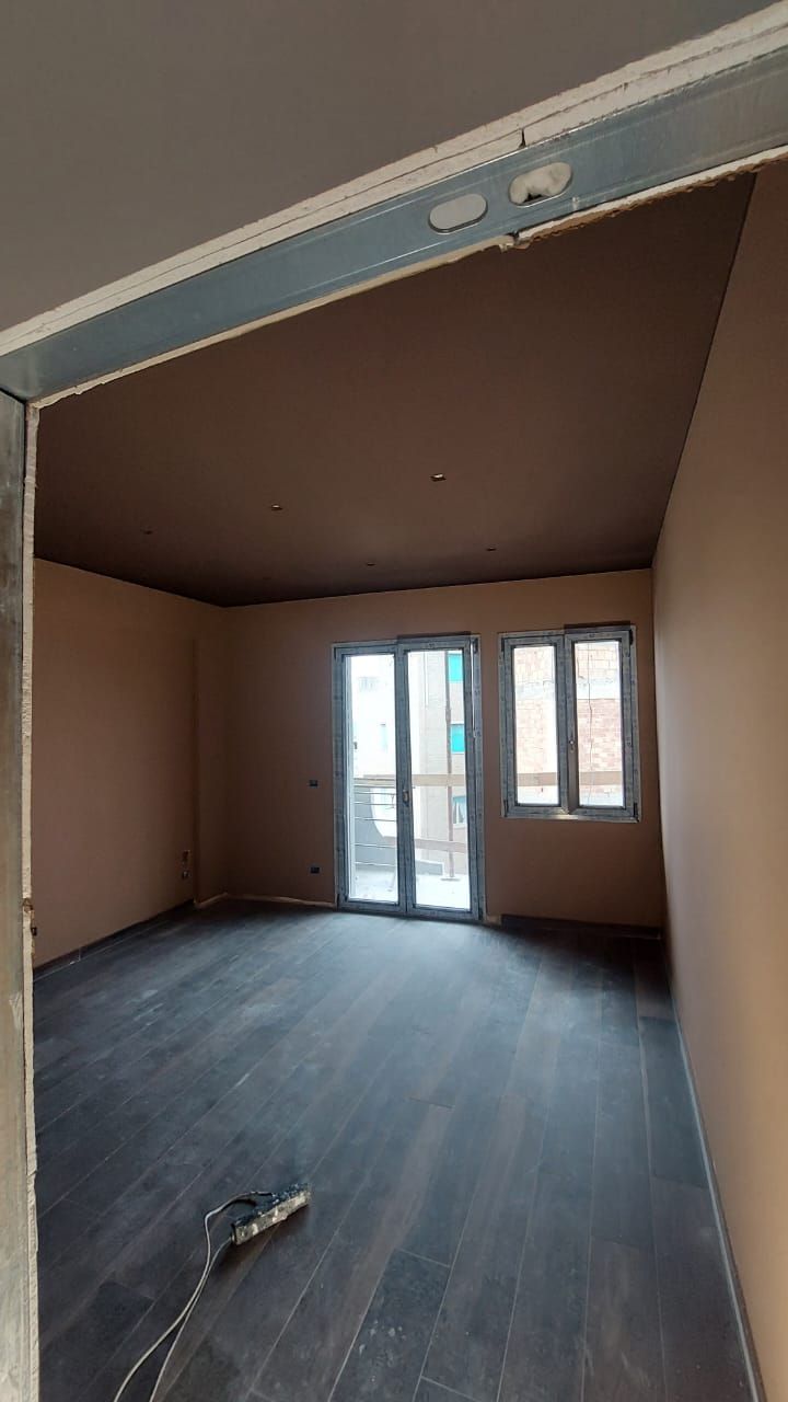 Stanza vuota con pareti e soffitto marrone chiaro, pavimento in legno scuro e porte a vetri che danno su un balcone.