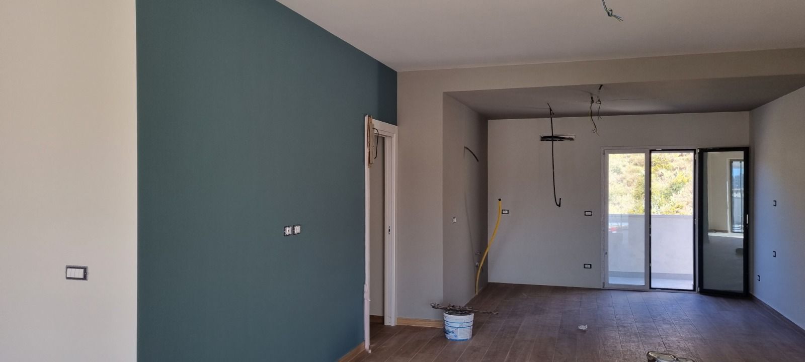 Una stanza incompiuta in fase di tinteggiatura. Una parete è verde acqua, le altre sono chiare. Ci sono fili che pendono dal soffitto.