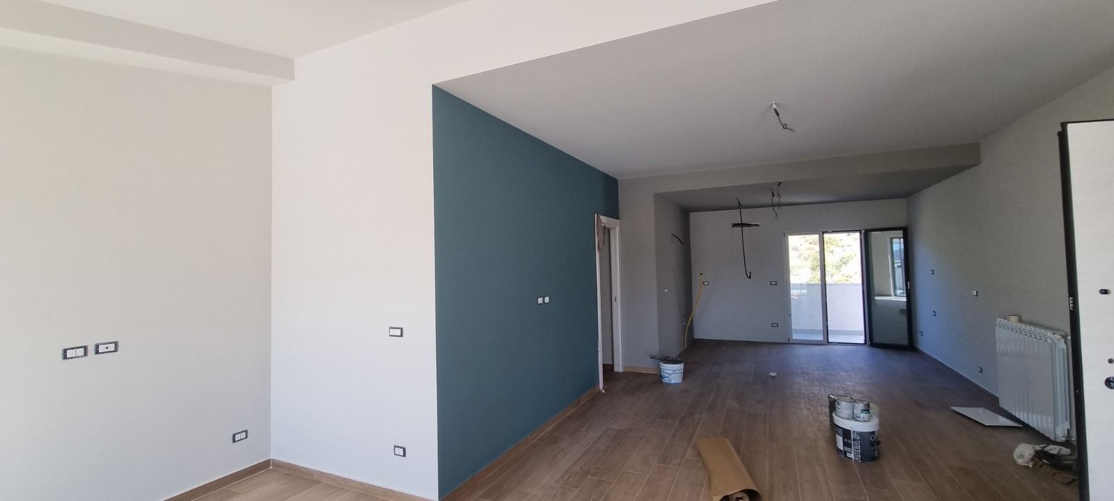 Interno di una stanza in costruzione con pareti bianche e verde acqua, pavimento in legno chiaro e balcone.