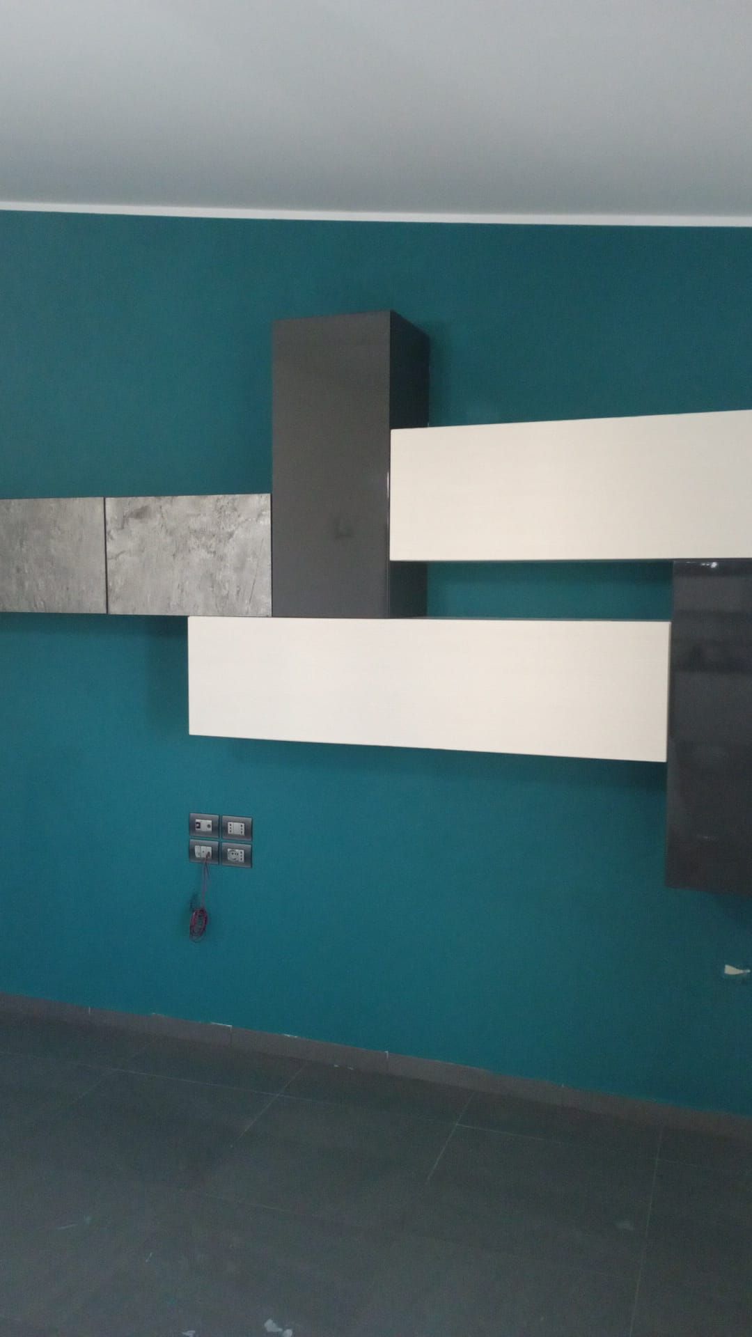 Parete verde acqua con motivo astratto: pannelli rettangolari bianchi, grigi e neri. Prese elettriche sotto.