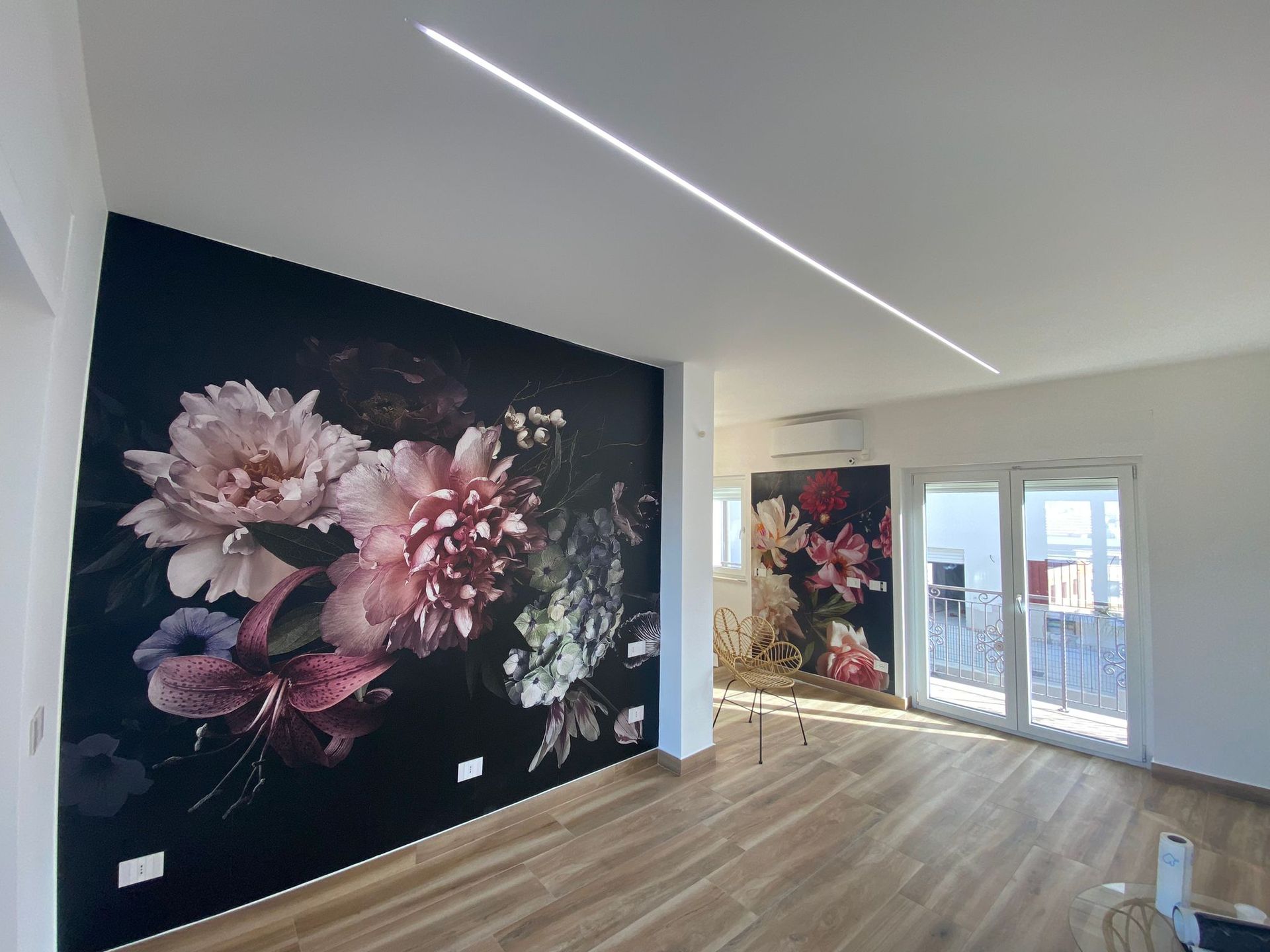 Camera con murale floreale e lampadario lineare sul soffitto. Pavimenti in legno, pareti bianche e porte-finestre.