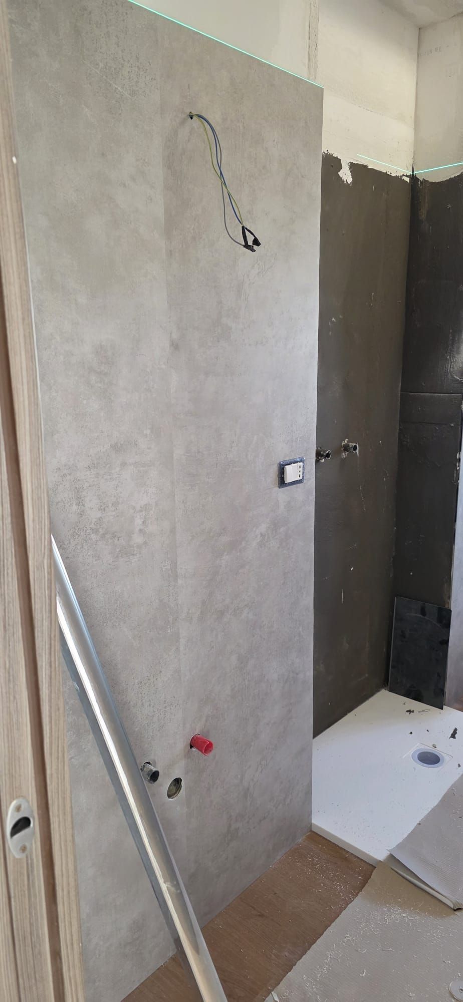 Bagno in costruzione. Pareti piastrellate grigie con tubature e cavi elettrici a vista. Pavimento bianco.