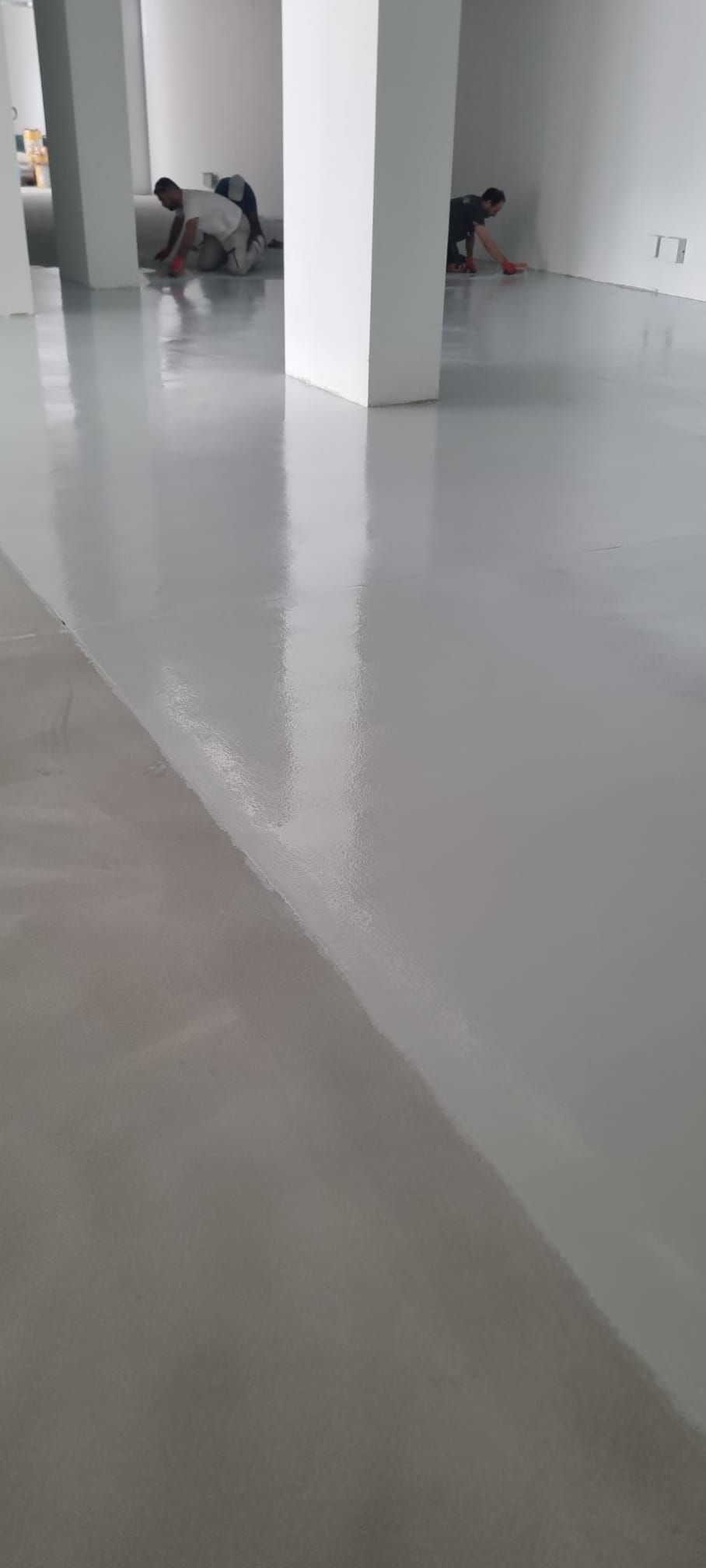 Operai che applicano la pavimentazione epossidica in un ampio spazio interno bianco. Il pavimento è di un colore grigio chiaro riflettente.