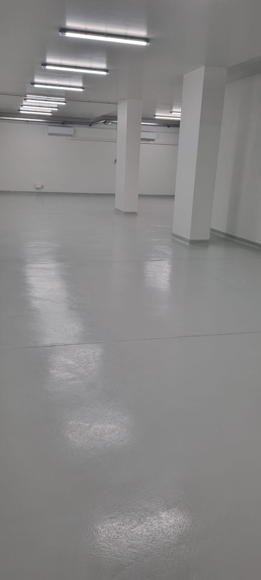 Stanza bianca, vuota e ben illuminata, con pavimento grigio riflettente e pilastri di sostegno.