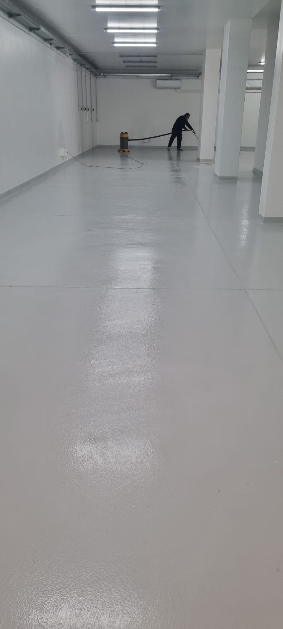 Persona che pulisce una stanza grande, vuota, con pareti bianche e un pavimento grigio chiaro e lucido.