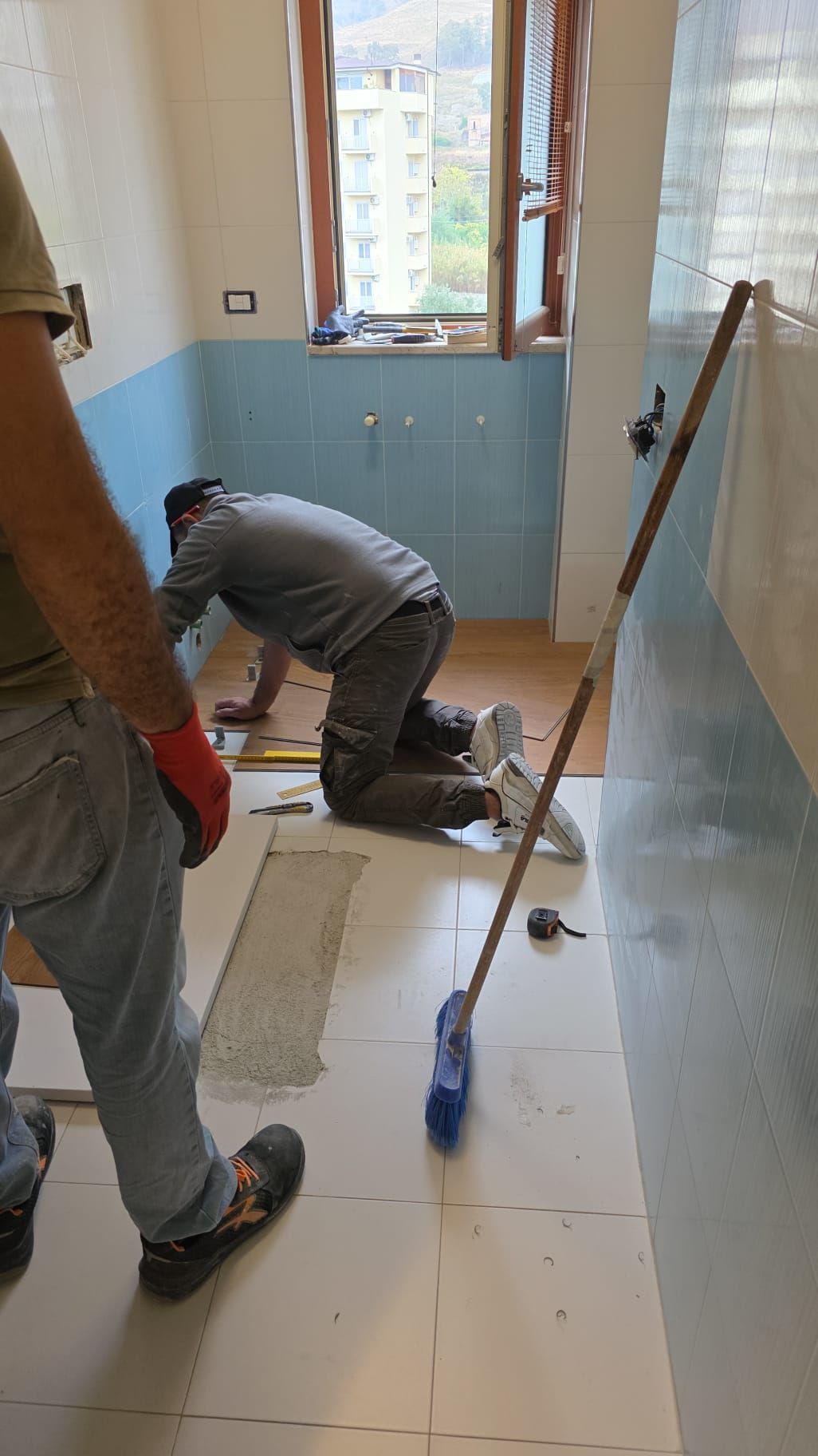 Due operai posano le piastrelle in un bagno, uno dei quali è inginocchiato. Pareti azzurre, pavimento effetto legno e finestra visibili.