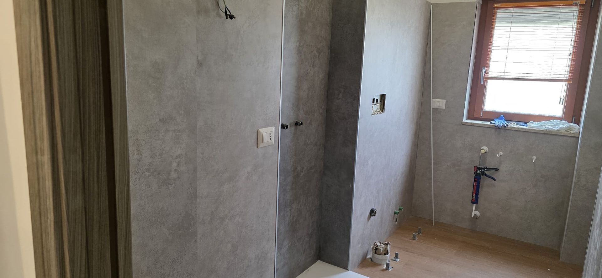 Bagno con pareti piastrellate grigie e finestra con persiane. Impianti idraulici a vista.