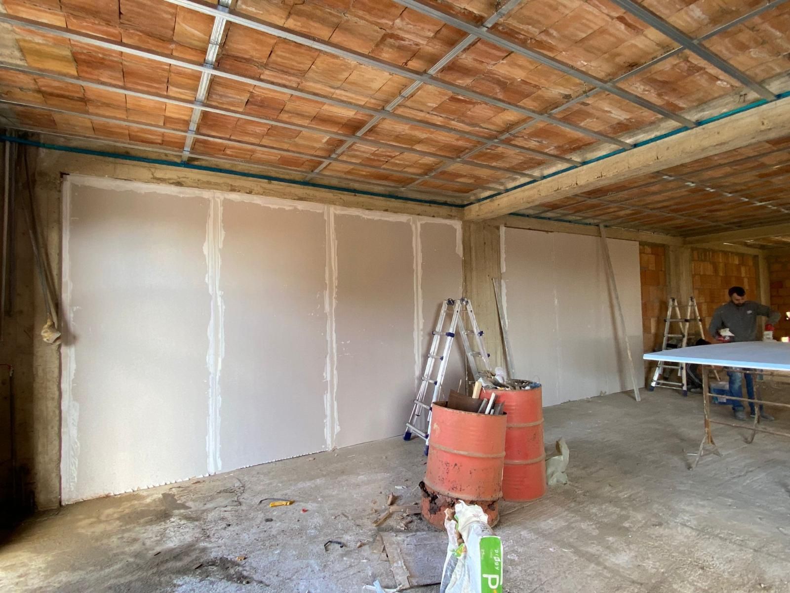 Cantiere edile interno. Cartongesso installato sulle pareti, scala, attrezzi e una persona che lavora sullo sfondo.