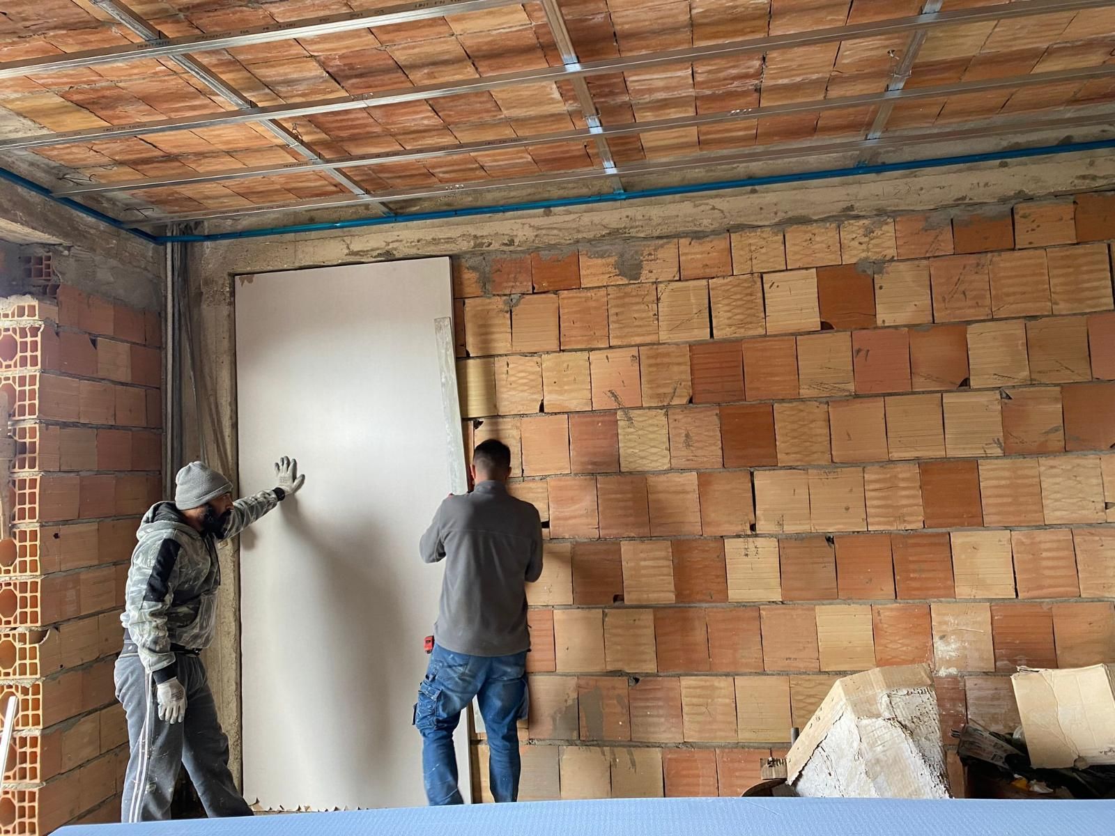 Due operai installano una lastra di cartongesso bianca su un muro di mattoni in un cantiere edile.