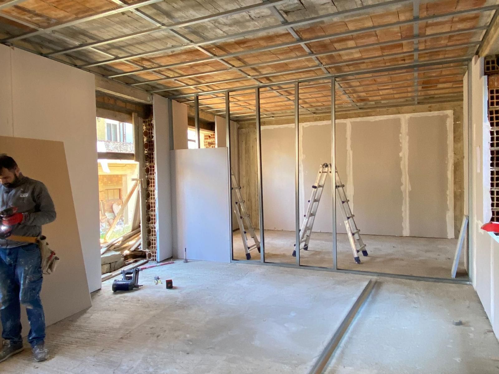 Interno di un cantiere, uomo al lavoro con cartongesso. Struttura in acciaio, pareti parzialmente rifinite, finestre aperte.