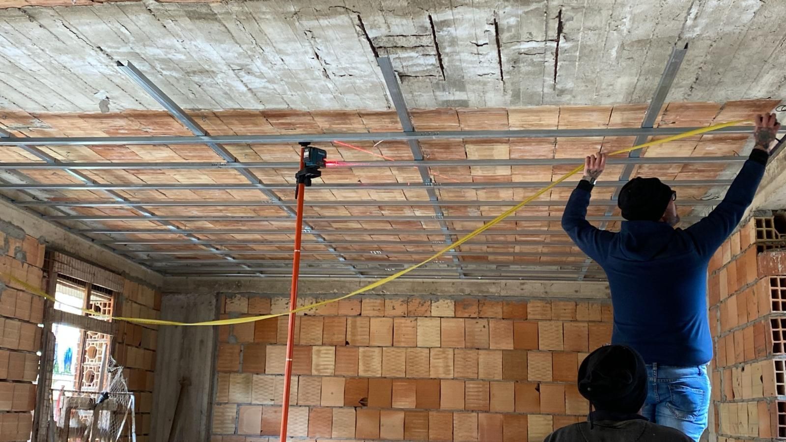 Uomo che installa un controsoffitto con griglia metallica in una stanza con pareti in mattoni. Filo elettrico giallo visibile.