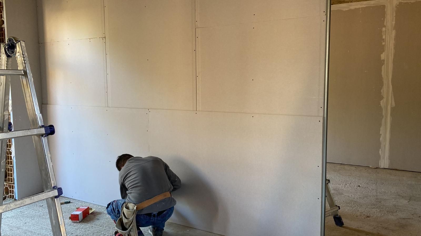 Persona che lavora su cartongesso, probabilmente in una stanza in costruzione, con indosso una maglietta grigia e jeans. Una scala è nelle vicinanze.