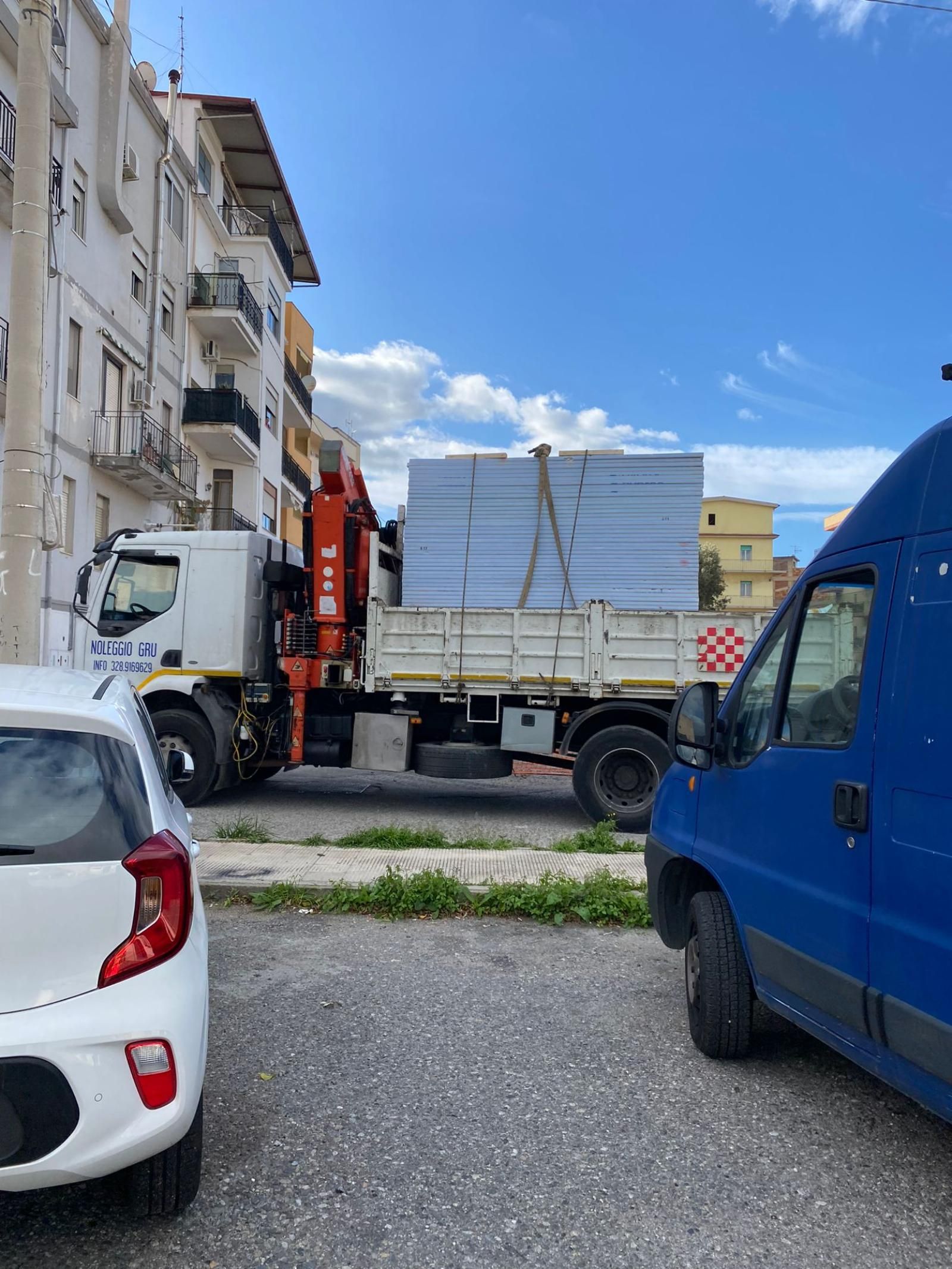Un camion bianco con una gru carica di pannelli rettangolari impilati, parcheggiato tra un'auto bianca e un furgone blu. Cielo azzurro.