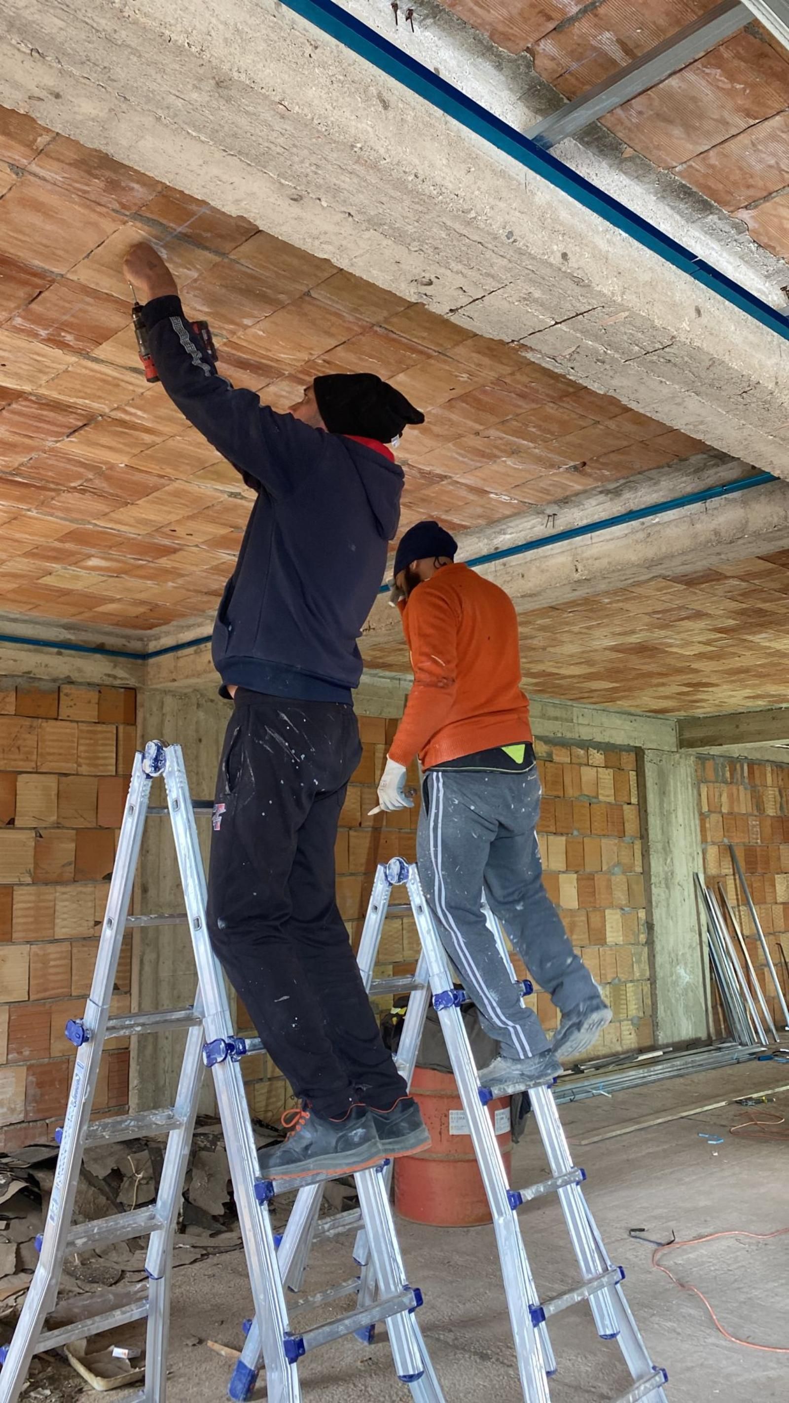 Due persone su una scala lavorano su un soffitto di un edificio. Una è protesa verso l'alto, l'altra è rivolta in avanti.