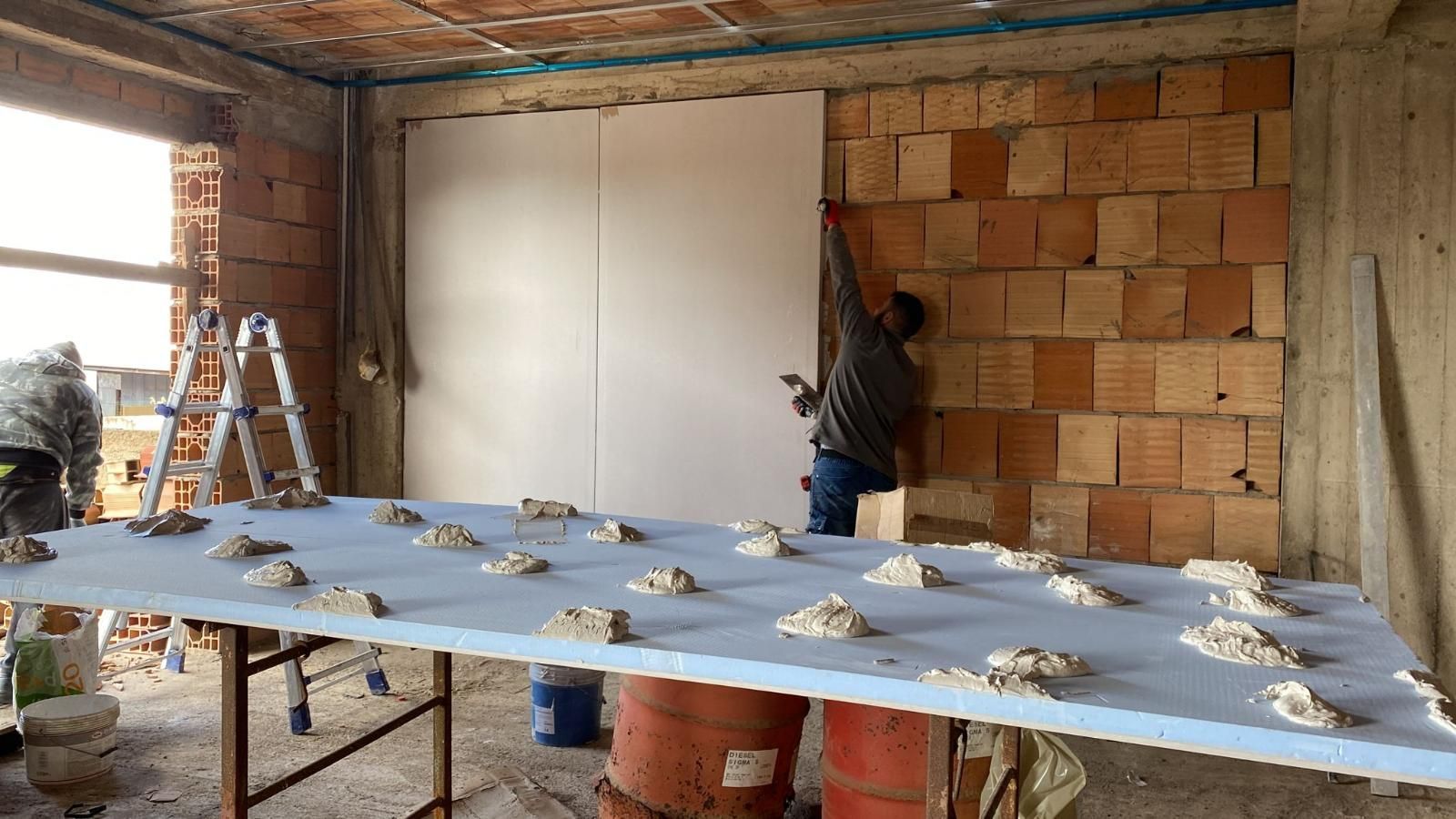 Persona installa pannelli in cartongesso su un muro di mattoni. In primo piano, un grande pannello di schiuma con adesivo. Cantiere edile.
