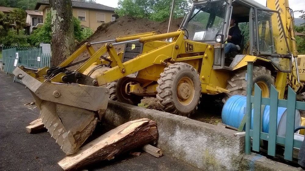 Una terna gialla spinge detriti in un cortile fangoso accanto a un muro di cemento e a una casa.