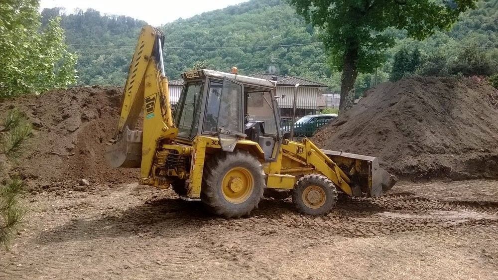 Una terna gialla in un cantiere sterrato con cumuli di terra e alberi sullo sfondo.