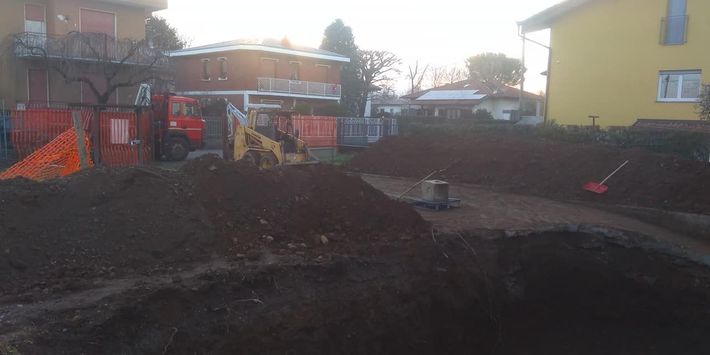 Cantiere edile con grandi cumuli di terra, barriere arancioni e case sullo sfondo.