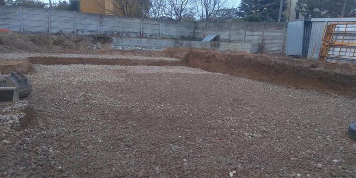 Cantiere edile riempito di ghiaia con bordi di terreno rialzati ed edifici sullo sfondo.