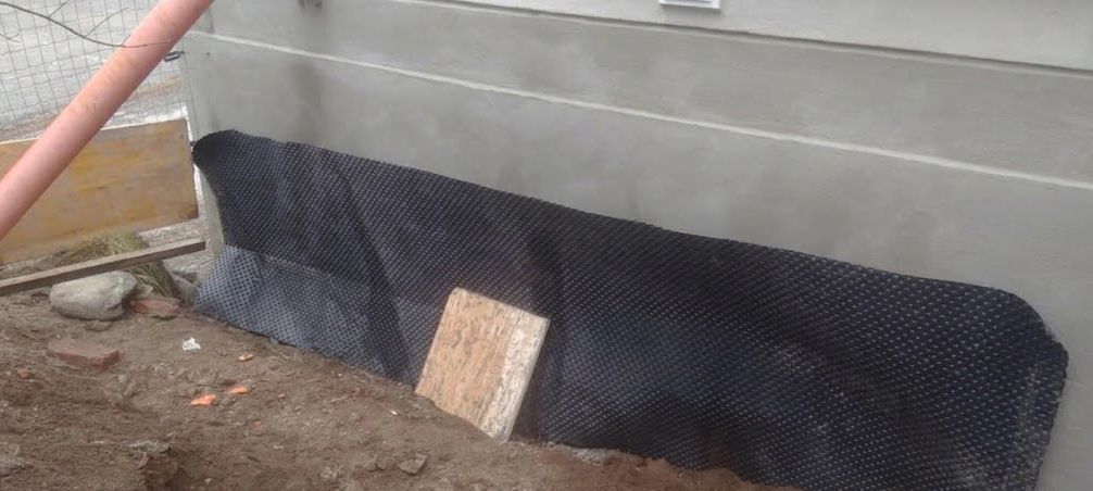 Una rete drenante nera ricopre una trincea di terra accanto a un muro di cemento, con un piccolo paletto di legno a fissarla.