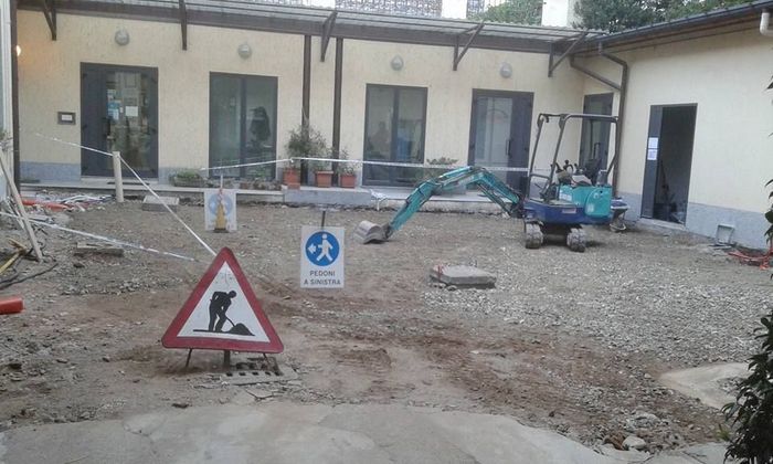 Cantiere edile in un cortile con attrezzi, cartelli e un piccolo escavatore vicino alle porte dell'edificio.