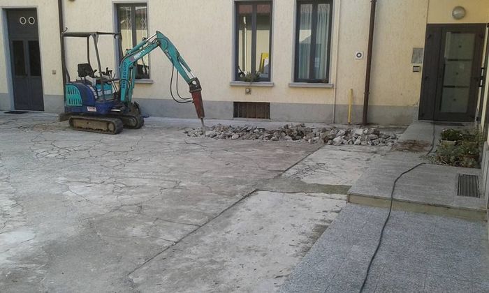 Un mini escavatore sta rimuovendo del cemento in un piccolo cortile accanto a un edificio.