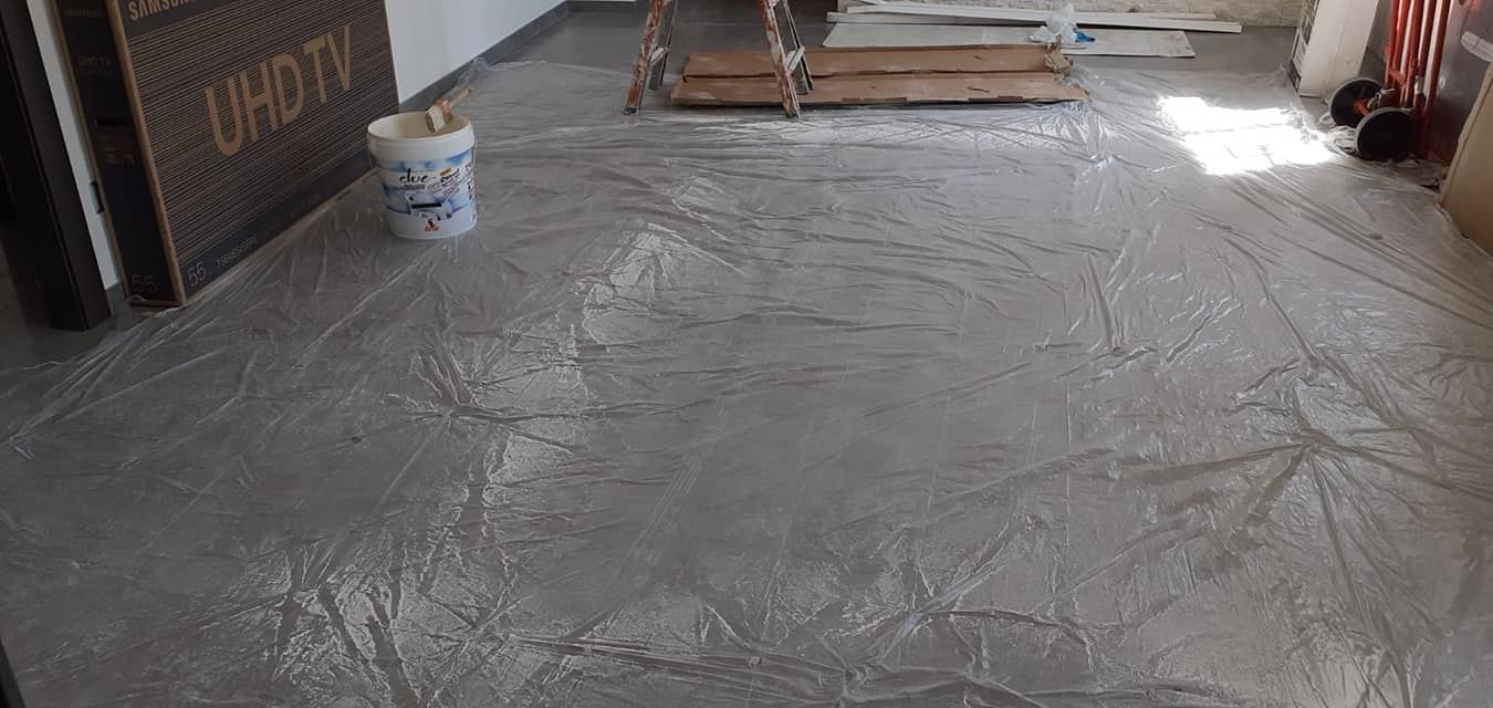 In una stanza interna, si sta lisciando un pavimento in cemento grigio, con un secchio e materiali edili nelle vicinanze.