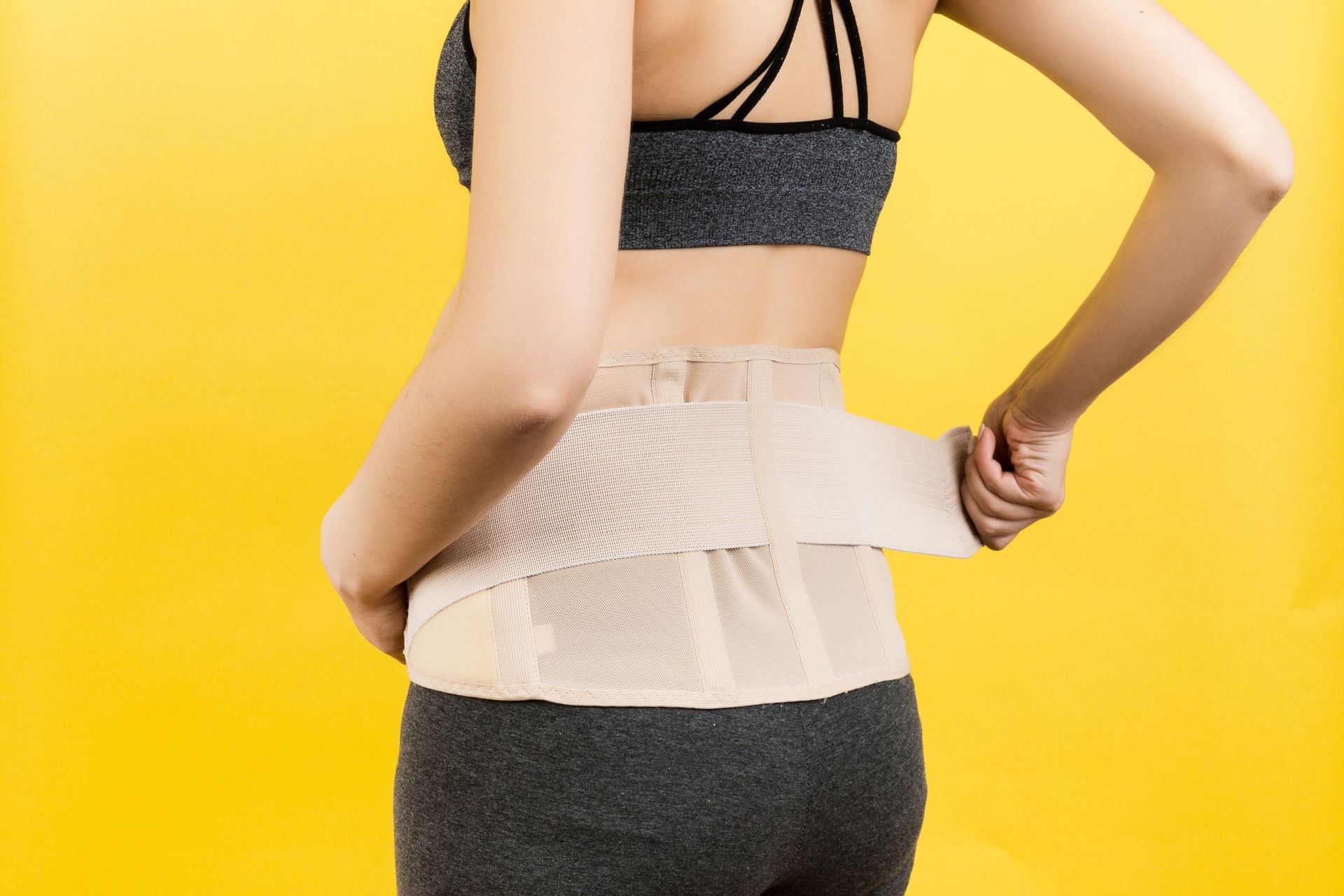 Woman putting on a back brace against a yellow backdrop. She wears a gray sports bra and pants.