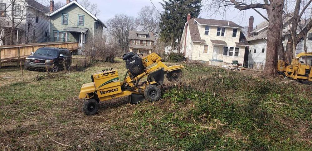 A yellow stump grinder is sitting in the middle of a grassy field in front of a house.