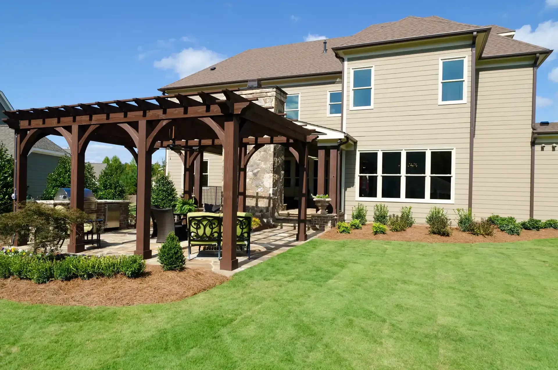 Brown pergola over patio with outdoor furniture, beside a beige house with blue sky.