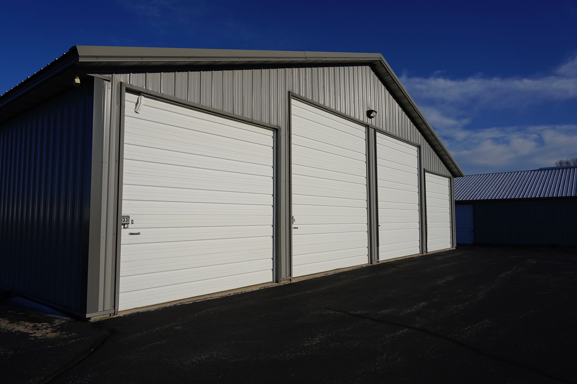 Three white garage doors on a gray metal building with dark pavement, under a blue sky.