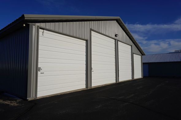 Three white garage doors on a gray metal building with dark pavement, under a blue sky.