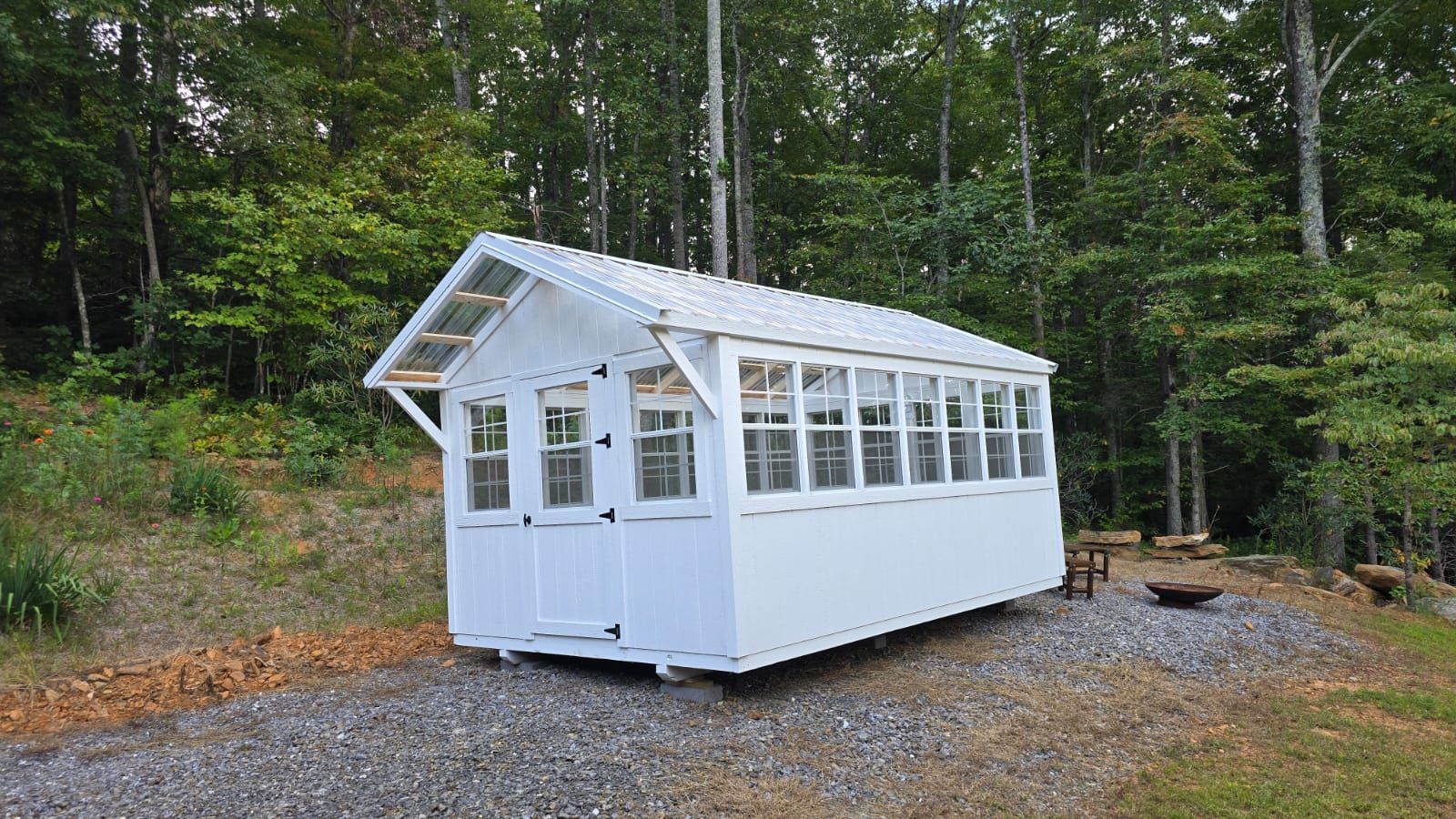 White greenhouse on a gravel pad in a wooded setting.
