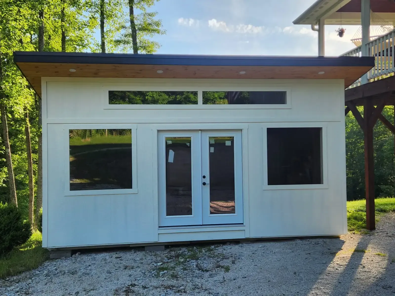 White shed with black trim, windows, and French doors. Gravel driveway, surrounded by trees.