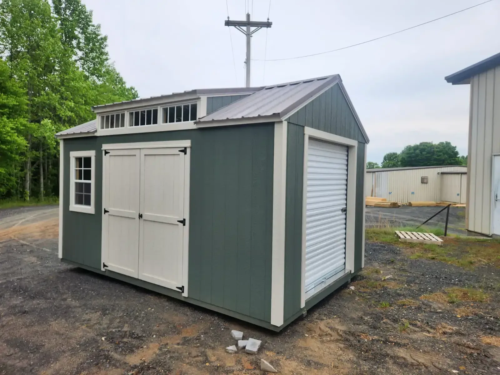 Green shed with white doors, window, and roll-up garage door; sits on gravel in front of a building.