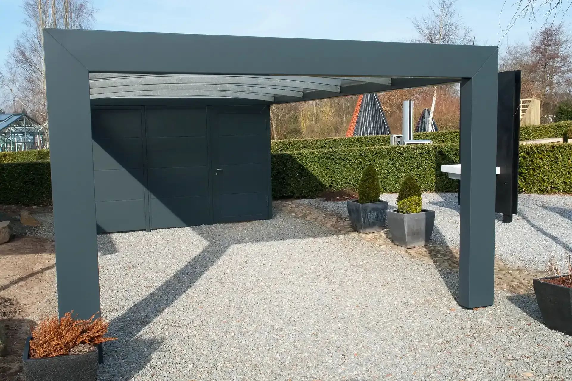 Grey-framed pergola with a dark grey storage unit, set on gravel with small trees in planters.
