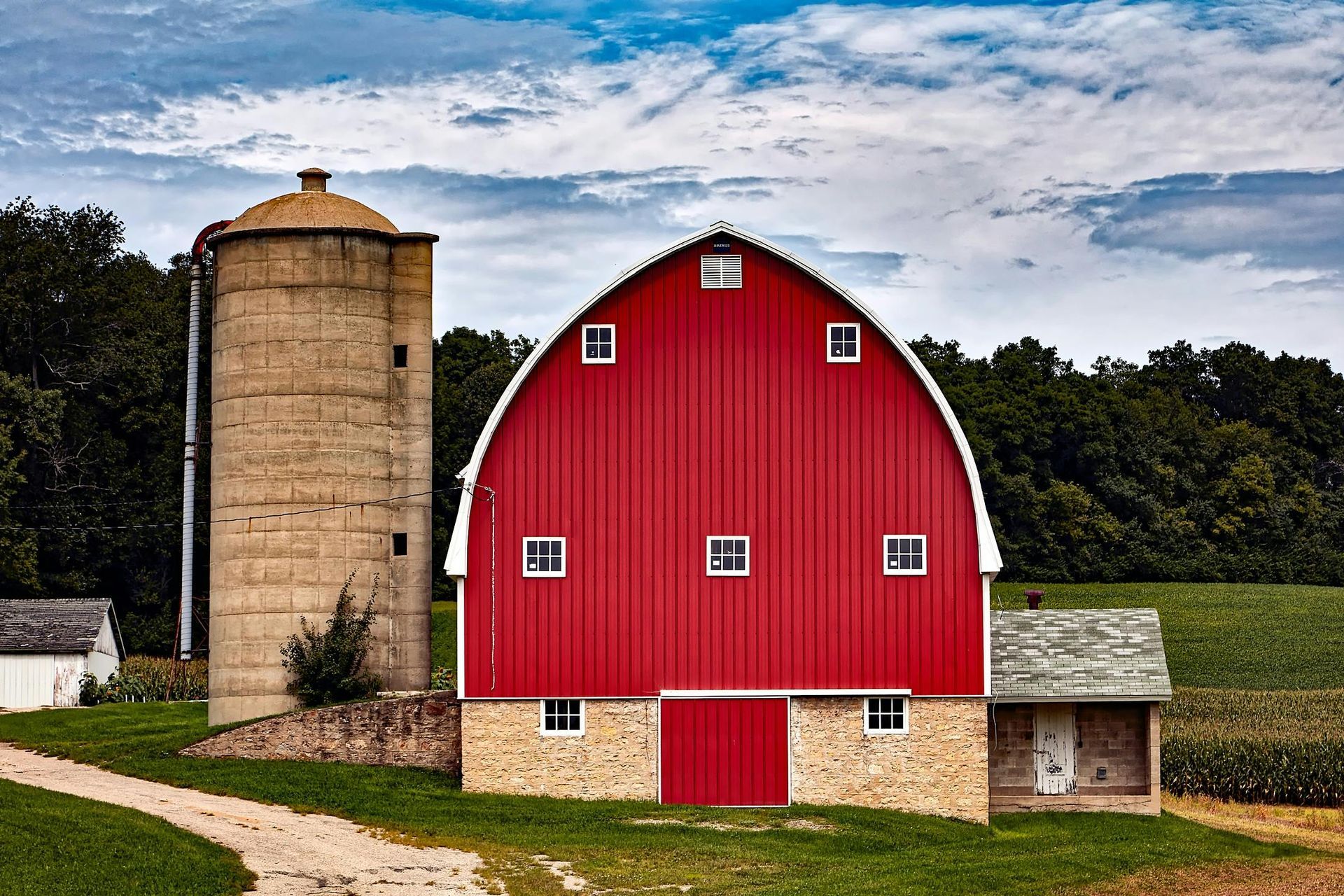 Red barn with silo in a rural setting, blue sky, stone foundation, and small building to the left.