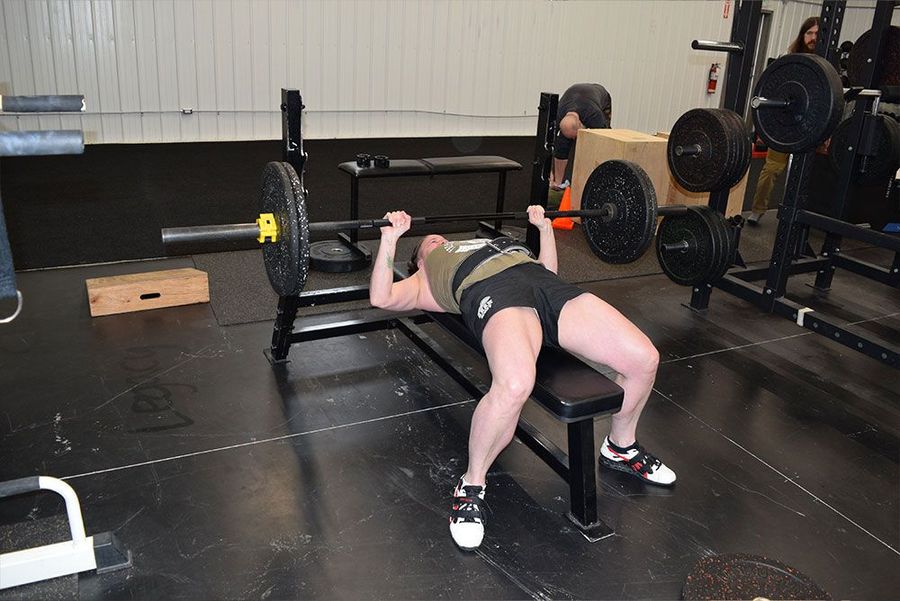 A person performs a barbell bench press exercise while lying on a flat weight bench inside a gym.