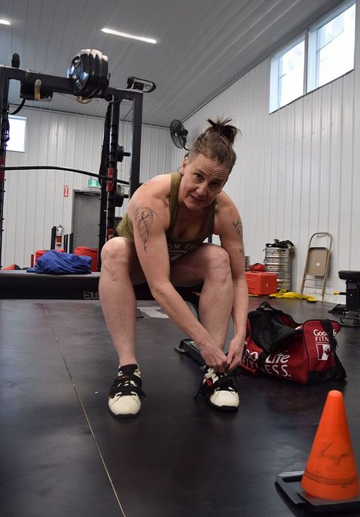A person in a gym squats to tie their white athletic shoes near a red gym bag and an orange cone.