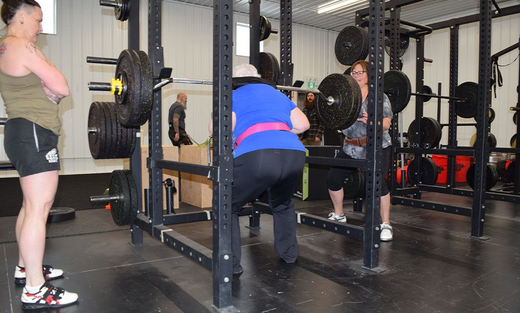 A weightlifter performs a squat inside a gym rack, supervised by an instructor, with another person spotting nearby.