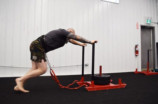 A person in a gym pushing a weighted red metal sled across a black floor.