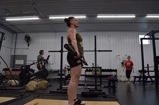 A person holding a barbell in a gym, standing in a neutral position with another individual walking in the background.