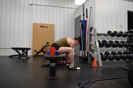 A person in an olive tank top sits on a gym bench, leaning forward with their head down in a bright, industrial-style gym.