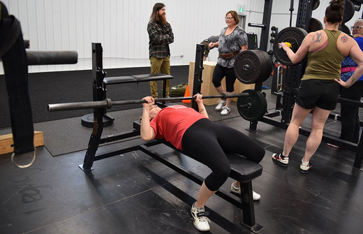 A person in a red shirt performs a barbell bench press in a gym, watched by three others standing nearby.