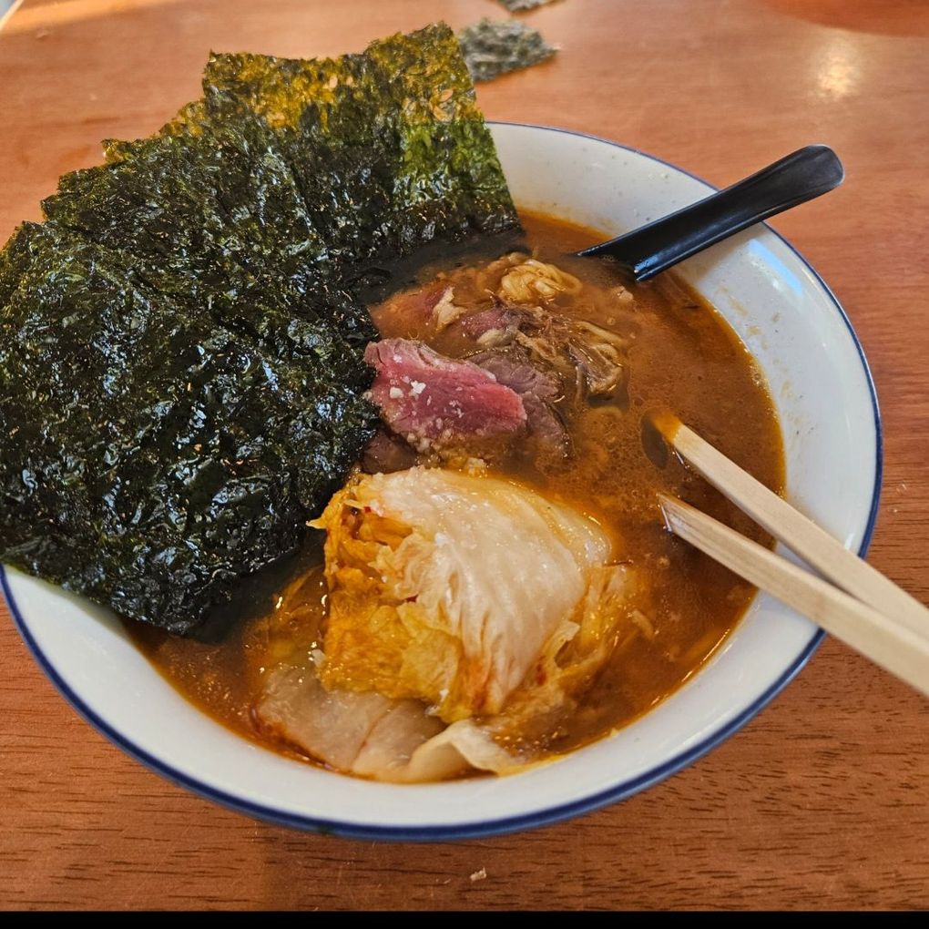 A bowl of ramen with dark broth, a large sheet of nori, kimchi, and a piece of raw red meat, served with chopsticks.
