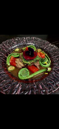 A gourmet dish featuring fiddlehead ferns, sautéed meat, and red tomatoes in a dark broth, garnished with a purple flower.
