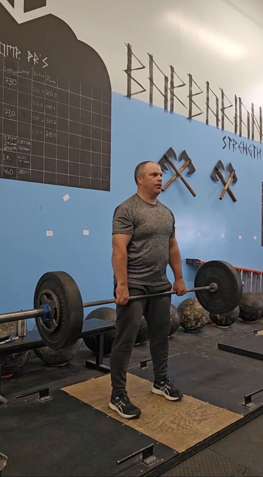 A person stands in a gym holding a barbell at thigh level during a deadlift in front of a wall with crossed axe art.