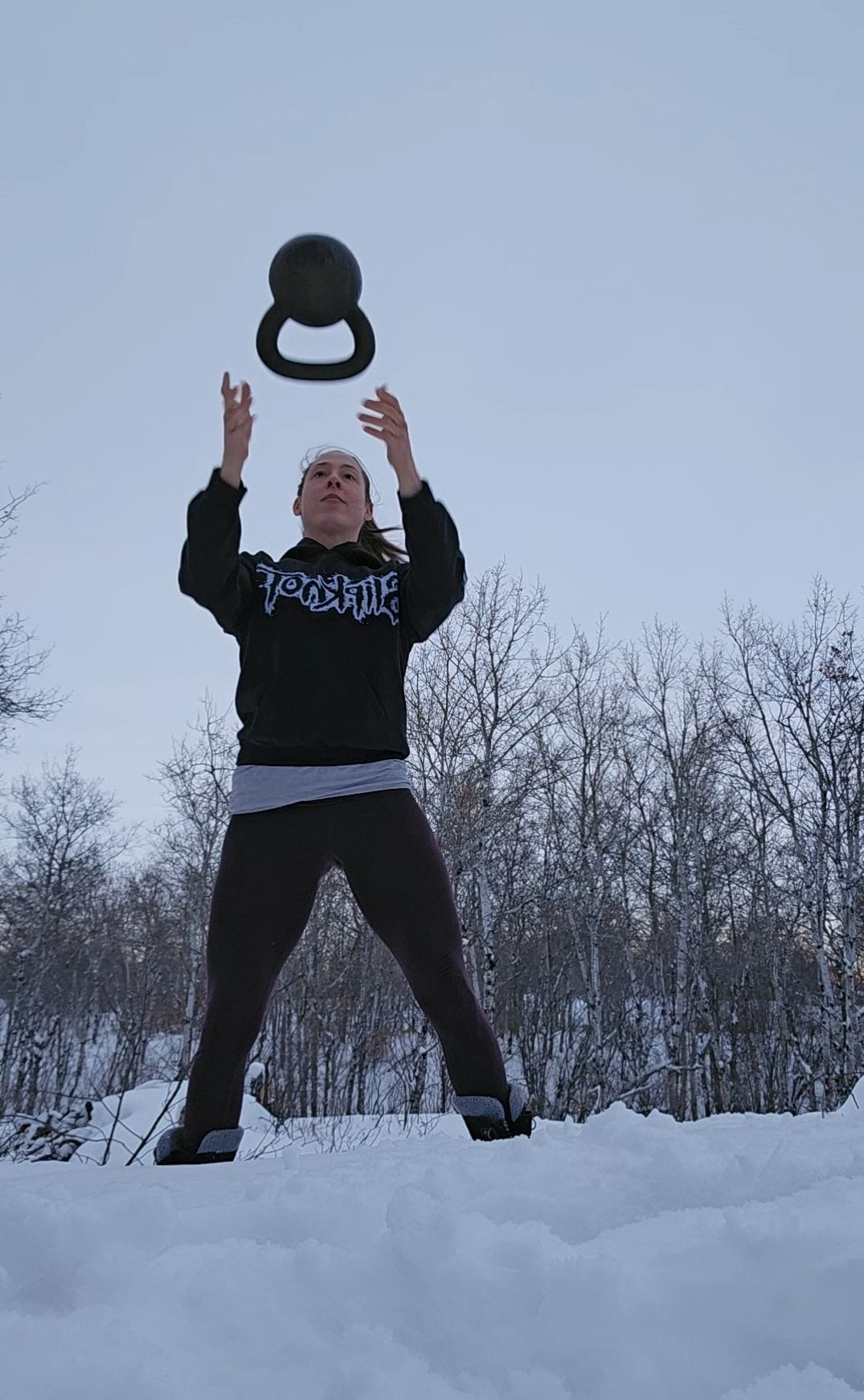 A person tosses a kettlebell into the air while standing in a snowy forest clearing.